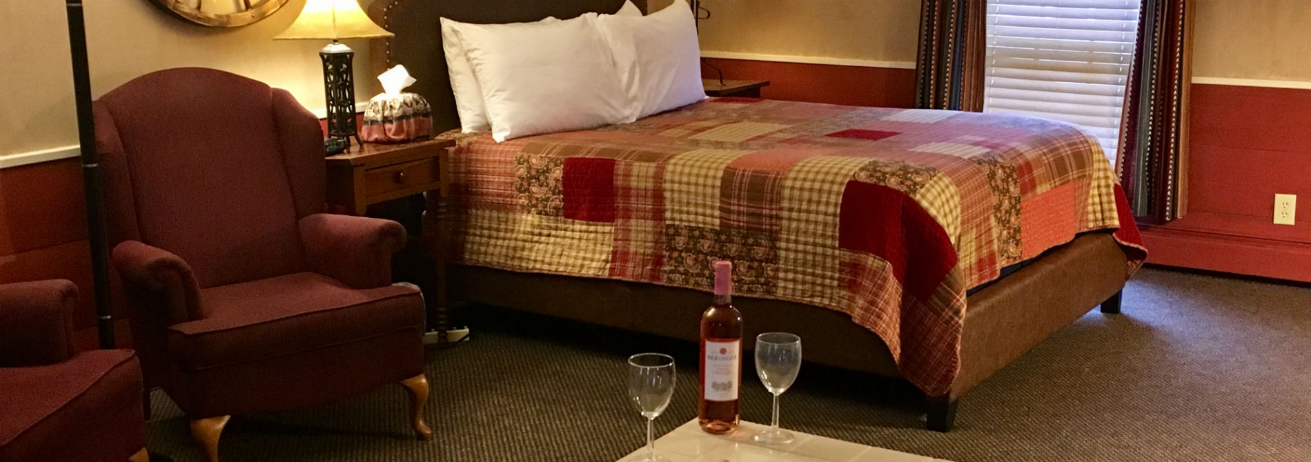 Patchwork quilt on a bed with pillows adjacent to a red chair with a bottle of wine in the foreground.
