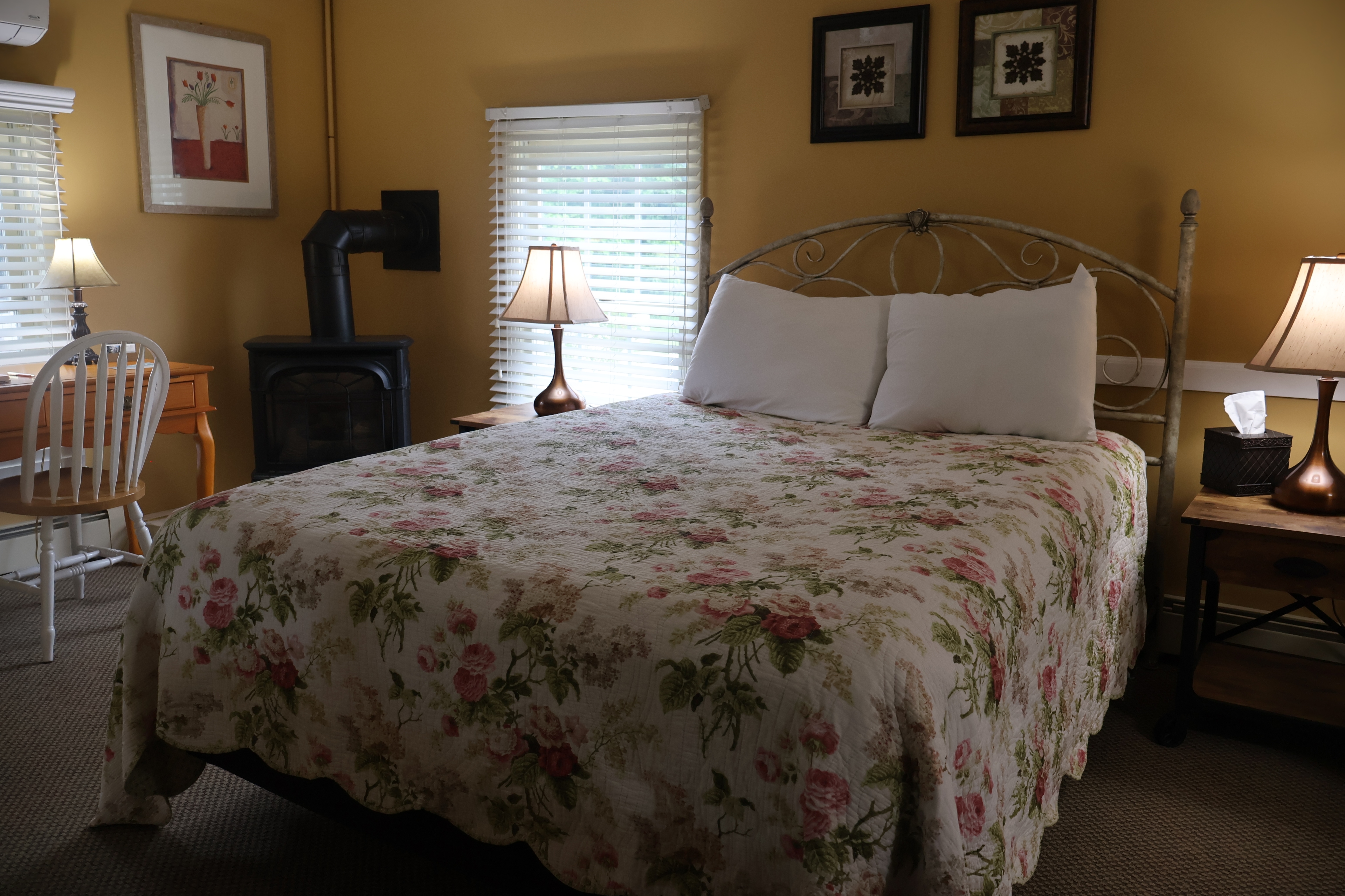 Queen bed with floral quilt in a mustard yellow room featuring a wood stove and desk.