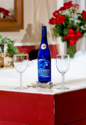 Blue bottle of wine with 2 wine glasses. Vase of roses in the background.