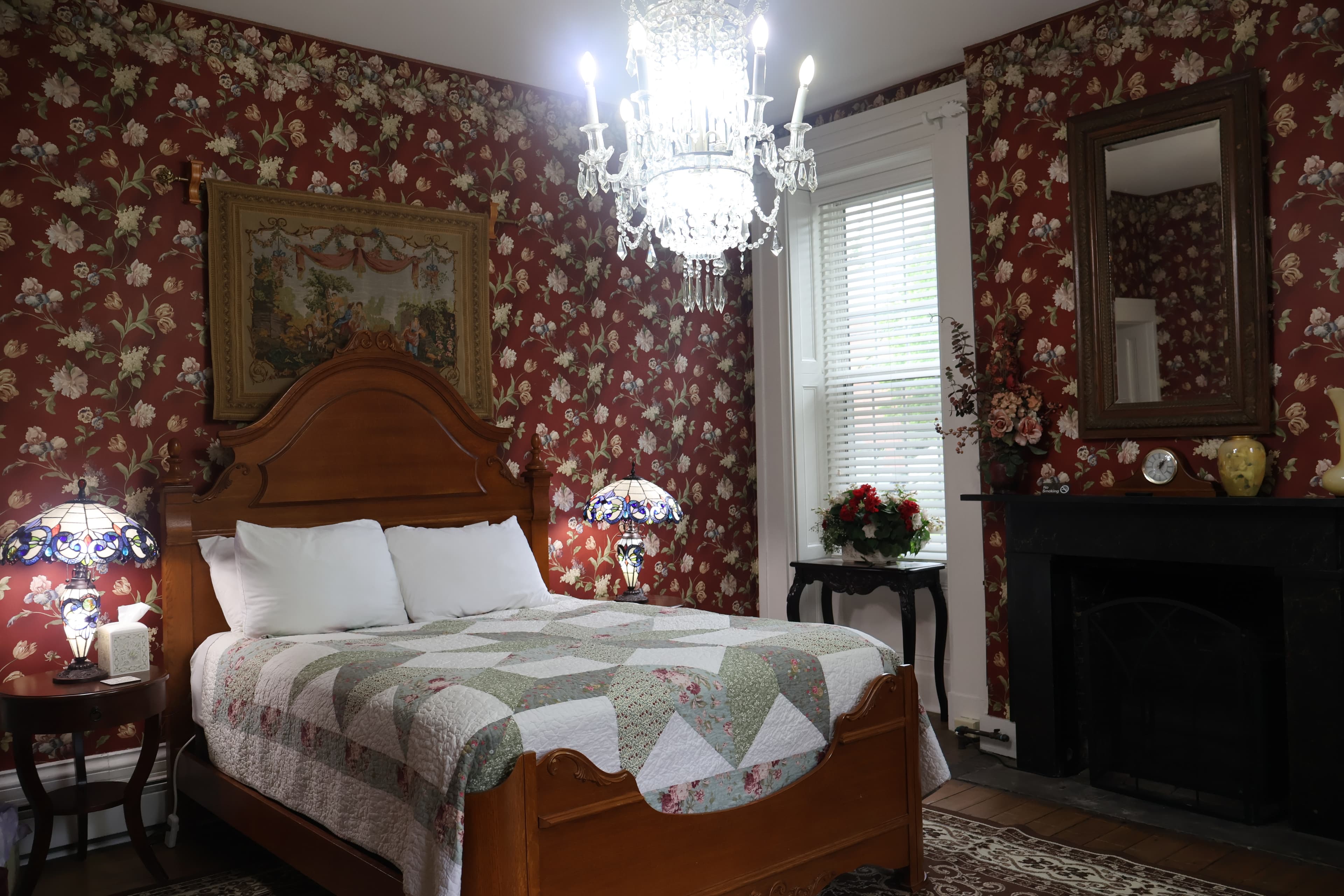 Victorian-style bedroom with red floral wallpaper, a wooden bed with a patchwork quilt, a black fireplace, and a crystal chandelier.