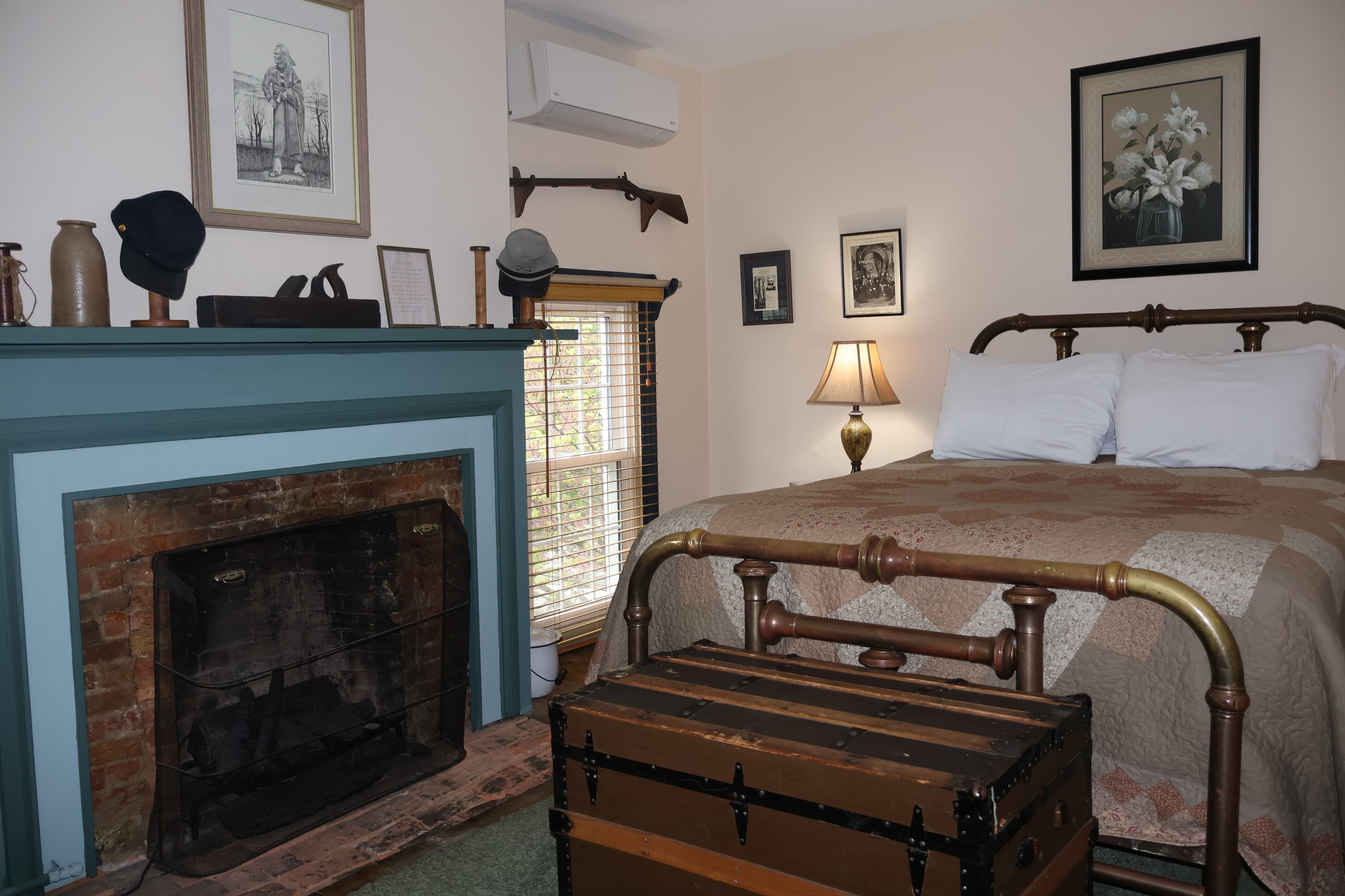 Bedroom with a brass bed, quilted brown bedspread, a teal-painted fireplace with exposed brick, and a vintage steamer trunk at the foot of the bed.