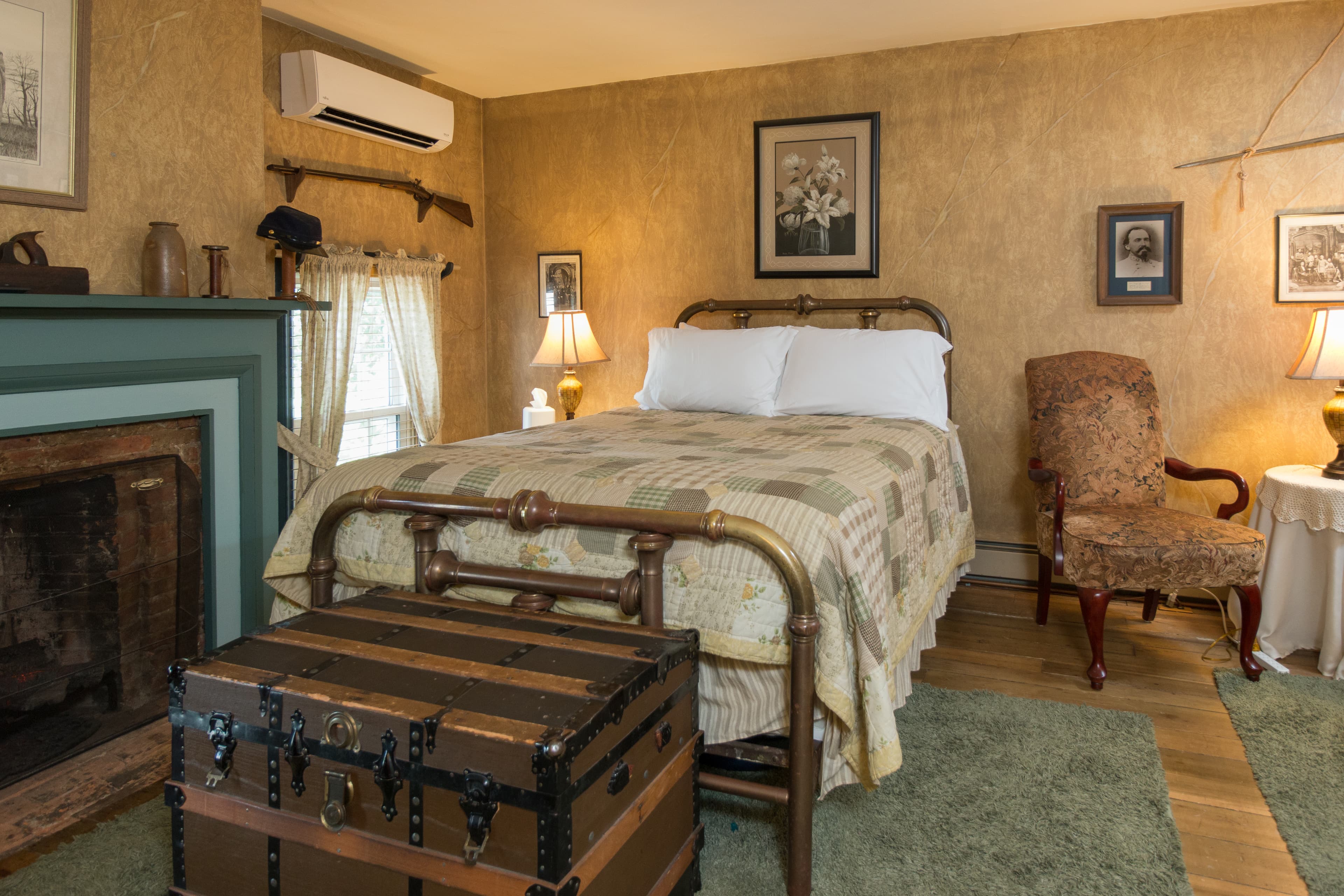 Bedroom with queen size metal bed frame topped with patchwork quilt, fireplace, vintage trunk, and cozy armchair next to an occasional table with lamp.