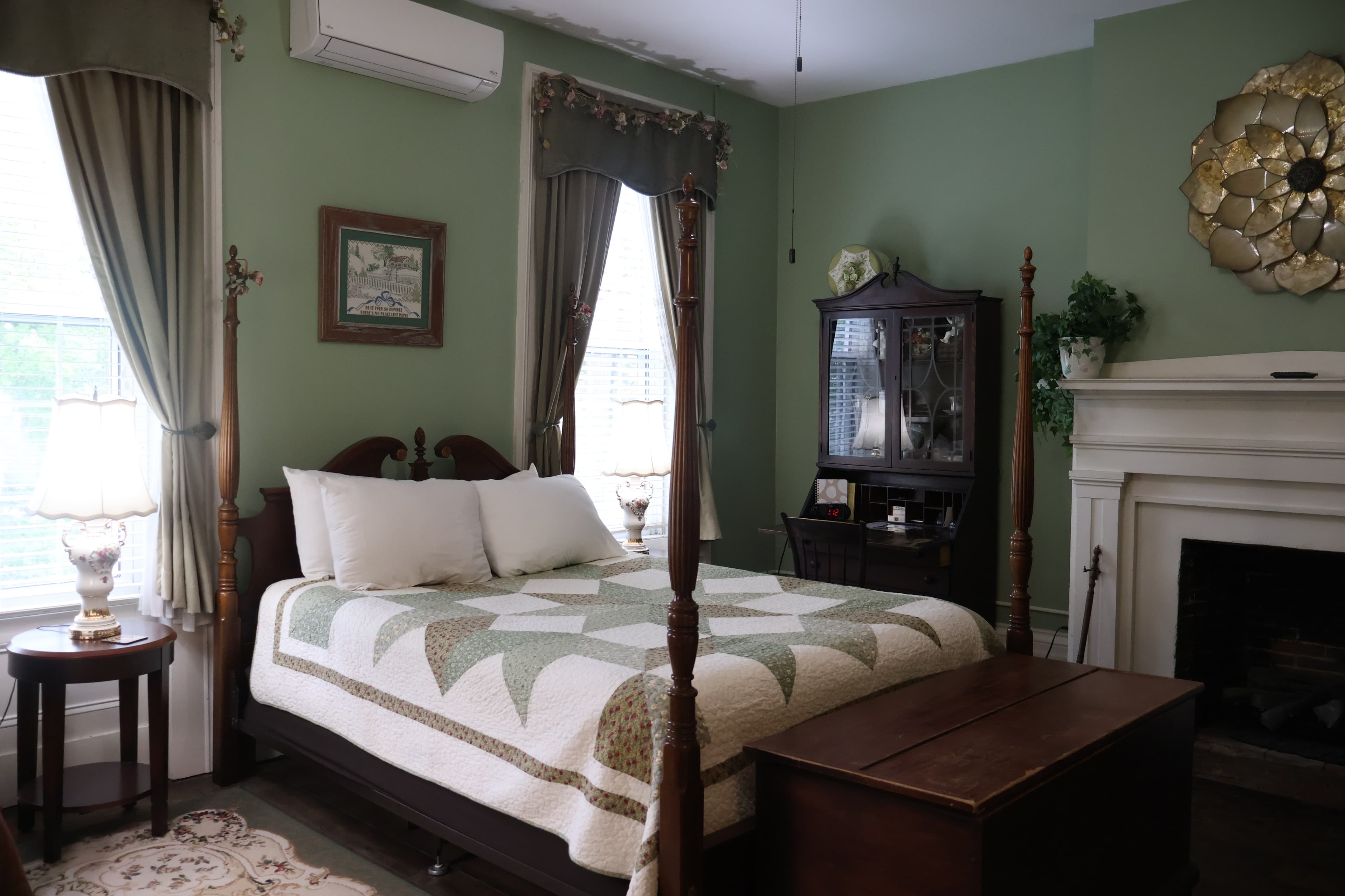 Queen bedroom with sage green walls, a wooden four-poster bed with a green and white quilt, a white fireplace, and a dark wood secretary desk.