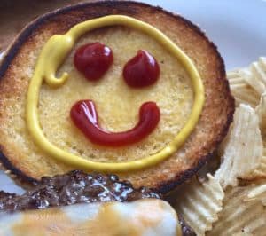 A toasted bun decorated with ketchup and mustard to resemble a smiling face, served with a burger and potato chips.