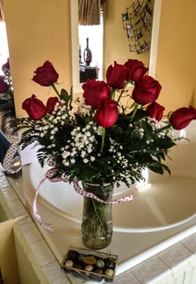 A bouquet of red roses with baby's breath in a glass vase, positioned beside a decorative box of chocolates on a counter.