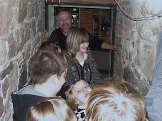 A group of children and adults gather near a narrow doorway, with one man smiling in the background.