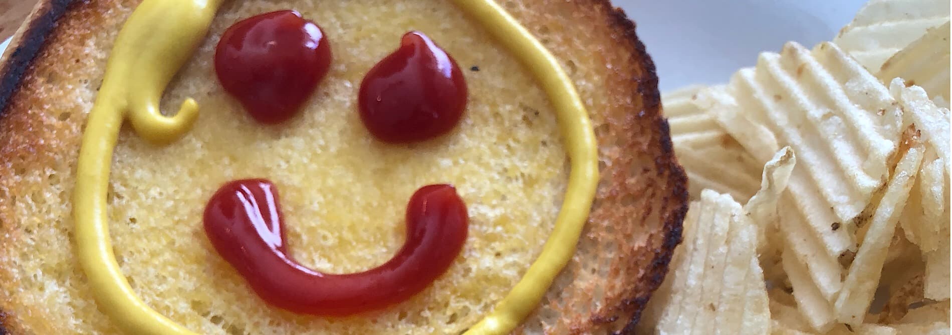 A slice of toast with a smiley face made of ketchup and mustard, accompanied by potato chips. A slice of toast with a smiley face made of ketchup and mustard, accompanied by potato chips.