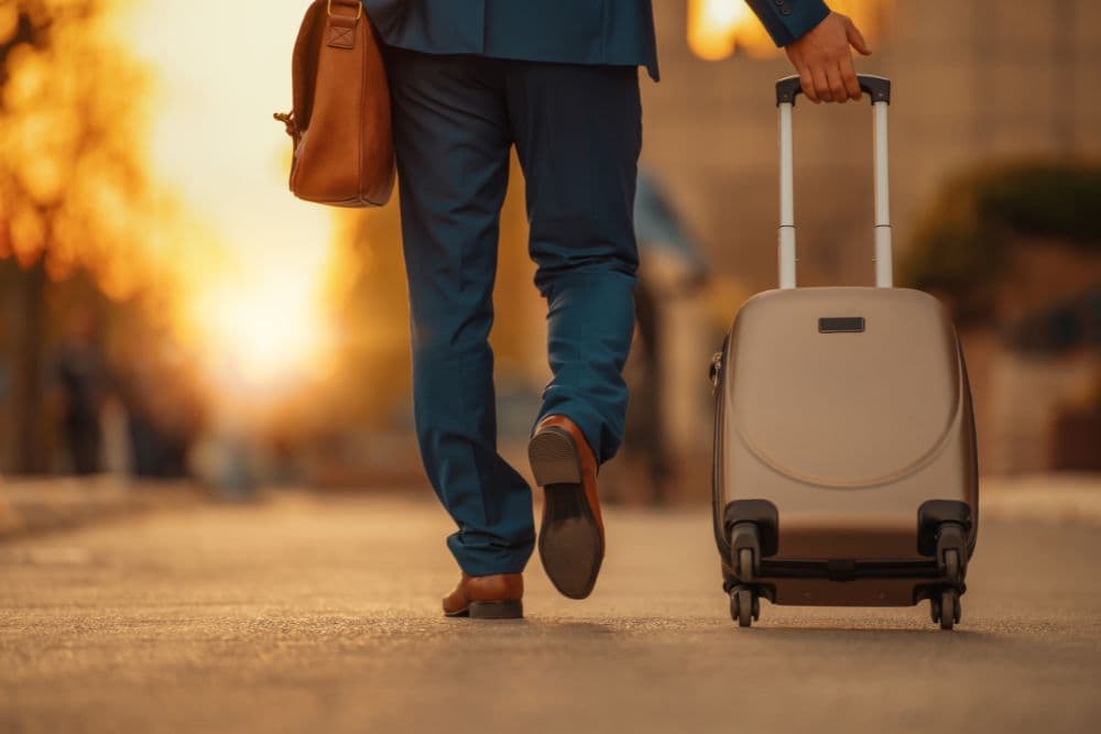 A man in a suit walks down a street, pulling a suitcase behind him as the sun sets.