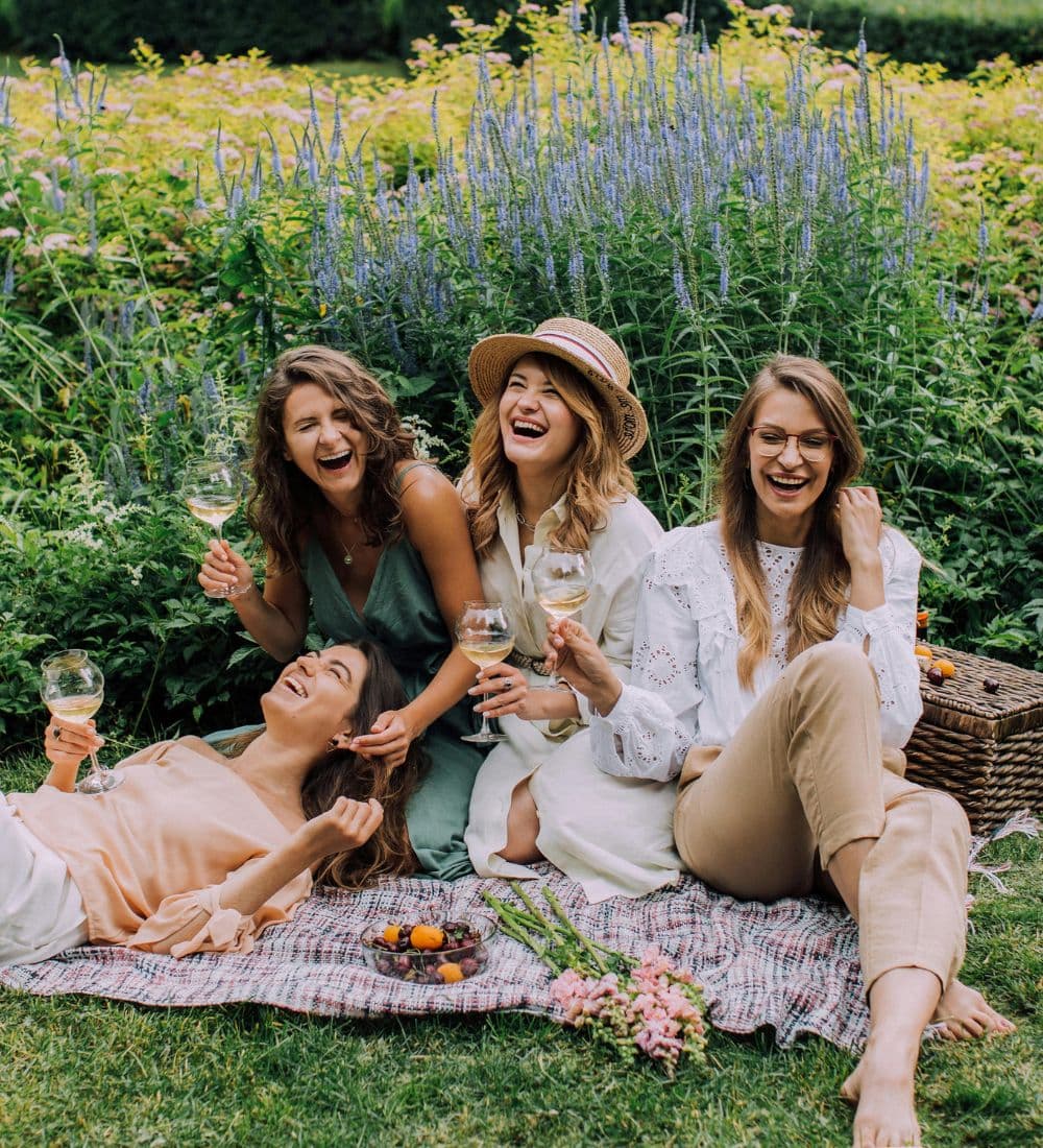 Four women laughing and enjoying drinks on a picnic blanket surrounded by vibrant flowers.