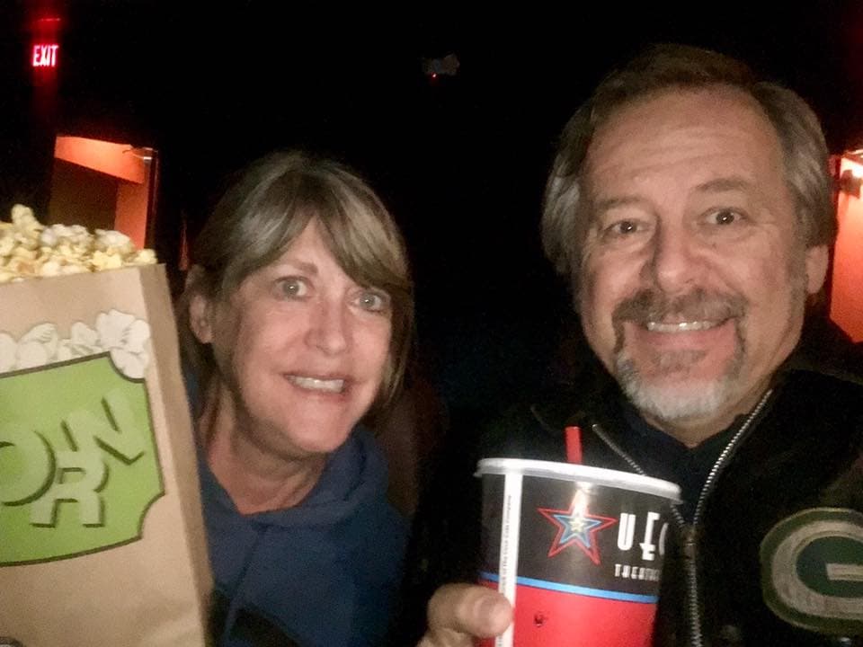 A man and woman smile while holding popcorn and a drink in a movie theater.