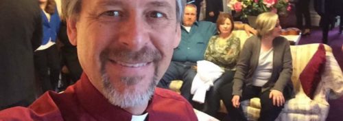 officiant in red shirt with religious collar in living room with people